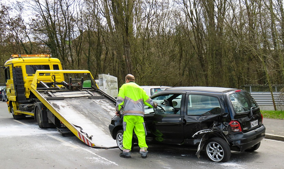 Photo of yellow flatbed towing truck about to tow damaged black car with employee standing in the side moving the steering wheel.